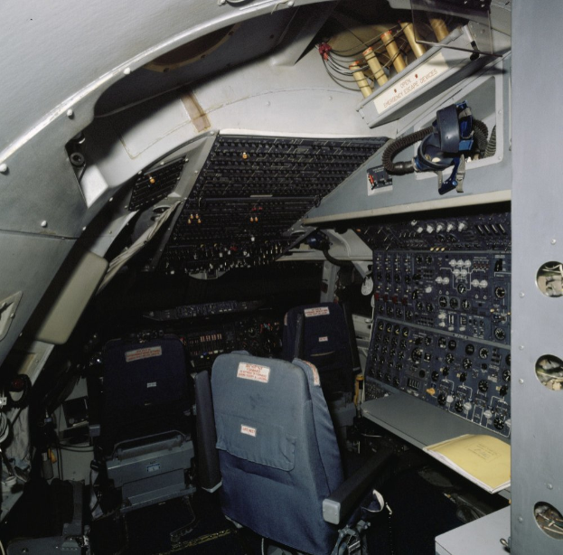 Das Cockpit eines Flugzeugs mit zwei Sitzen, einem Tisch mit einem Buch und verschiedenen elektronischen Geräten und Kabeln.