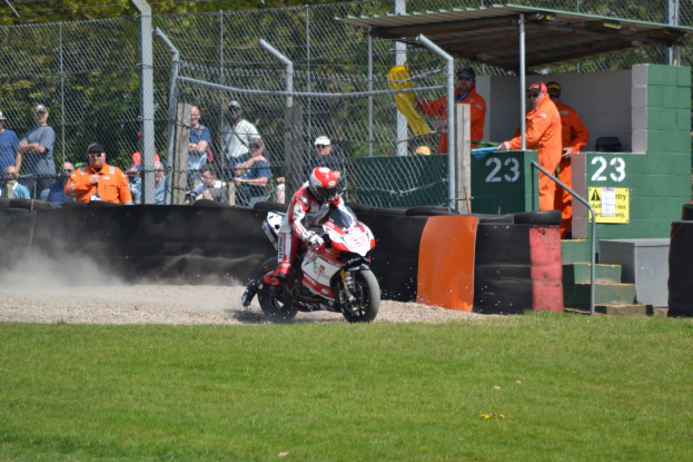 Eine Person in Helm und Handschuhen fährt ein Motorrad über ein grasbewachsenes Feld, mit Zuschauern in Mützen und Brillen, einem Zaun, Treppen mit Geländern und einem nummerierten Schuppen im Hintergrund.