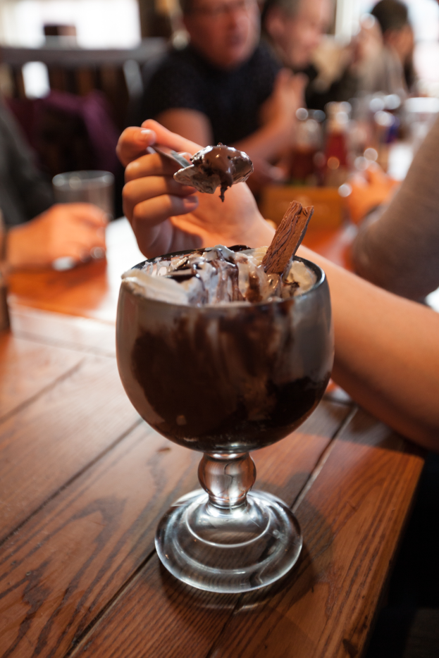 Eine Gruppe von Menschen sitzt um einen Tisch mit einem Glas Schokoladenpudding in der Mitte, eine Person im Vordergrund isst den Pudding mit einem Löffel, Hintergrund leicht unscharf.