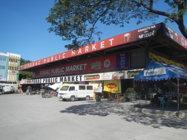 Öffentlicher Markt mit einem vor ihm geparkten Van, umgeben von Gebäuden, Geschäften, Namensschildern, Kraftfahrzeugen, Menschen unter einem Zelt sitzend, Geländern, Bäumen und einem klaren blauen Himmel.