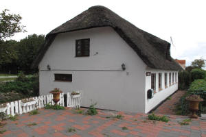 Ein weißes Haus mit einem Reetdach und einem Backsteinweg, umgeben von einem weißen Holzzaun, mit Fenstern, Topfpflanzen, Gras, einer Gruppe von Bäumen und einem bewölkten Himmel im Hintergrund.