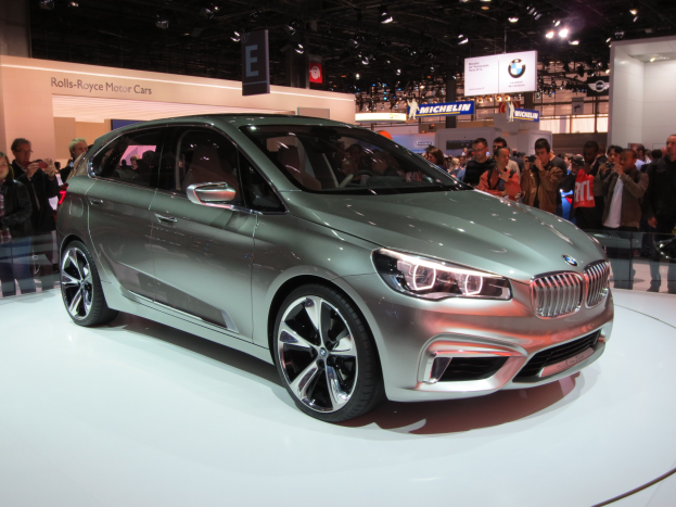 Ein BMW 2er Active Tourer Concept-Fahrzeug, das auf der Frankfurter Motor Show ausgestellt wird, umgeben von Menschen, die Fotos machen, unter Deckenlampen mit Texttafeln im Hintergrund.