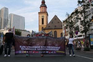 Eine Gruppe maskierter Menschen marschiert mit einem Transparent die Straße entlang, mit einem geparkten Auto auf der linken Seite, Gebäuden, Bäumen, Schildern, Polen und einem Uhrenturm im Hintergrund unter einem klaren blauen Himmel.