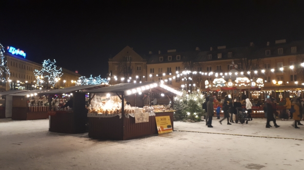 Lebendiger Weihnachtsmarkt an einem verschneiten Abend mit Menschen, Buden mit Lichtern, Pflanzen, Bäumen, Gebäuden, Schildern und einem bewölkten Himmel.