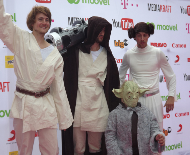 Eine Gruppe von Menschen in Star-Wars-Kostümen, die auf einem roten Teppich posieren, wobei einer ein Objekt und ein Banner mit Text und Logos im Hintergrund hält.