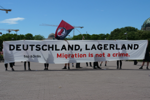 Eine Gruppe von Menschen hält ein Transparent mit der Aufschrift "Deutschland, Lagerland Migration ist kein Verbrechen" und eine Flagge, steht auf dem Boden mit Straßenlaternen, Bäumen, Kränen, einem Gebäude und einem bewölkten Himmel im Hintergrund.