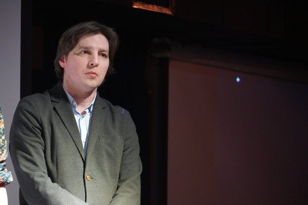 Zwei Personen stehen nebeneinander auf einer Bühne, der Mann im Anzug und die Frau neben ihm, beide mit ernsten Gesichtern.