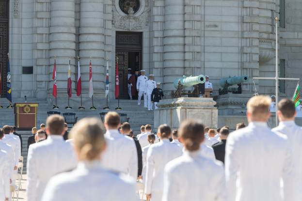 Gruppe von Menschen in weißen Marineuniformen vor einem Gebäude mit Säulen, einem Podium und Treppen während einer Abschlussfeier, mit Fahnen, Mikrofon und Kanonen im Hintergrund.