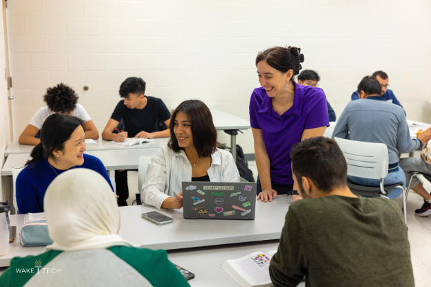 Eine Gruppe von Menschen in einem Klassenzimmer sitzt um einen Tisch und diskutiert über verschiedene Gegenstände wie Laptops, Bücher, Handys und Taschen, wobei eine Tür im Hintergrund zu sehen ist.