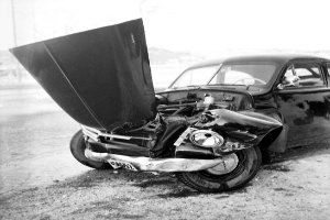 Ein Schwarz-Weiß-Foto eines schwerbeschädigten Autos am Straßenrand mit eingedellter Motorhaube, zersplitterten Fenstern und geöffneter Fahrertür, die den Motor freigibt.