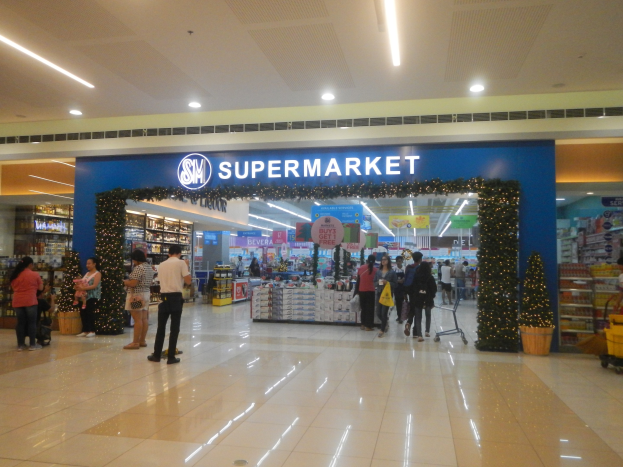 Menschen vor einem Supermarkt mit Einkaufstaschen, einigen mit Einkaufswagen, Topfpflanze, Regalen, Schildern und Deckenbeleuchtung.