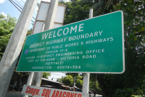 Ein grünes Schild mit der Aufschrift 'Willkommen an der Bezirksgrenze' ist an zwei weißen Pfählen befestigt, mit Fahrzeugen, Gebäuden und Bäumen im Hintergrund unter einem blauen und weißen Himmel.