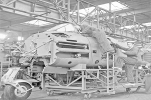 Ein Schwarz-Weiß-Bild einer Gruppe von Männern, die an einem Flugzeug in einer Fabrik arbeiten, mit anderen Fahrzeugen, Metallrahmen und einer Decke mit Beleuchtung.