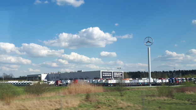 Ein Mercedes-Benz-Werk in Deutschland mit Fahrzeugen auf der Stra├če, umgeben von Gras, Pflanzen, B├Ąumen, Geb├Ąuden und Masten, darunter einer mit einem Logo, unter einem bew├Âlktem Himmel.
