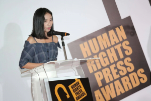 Eine Frau in einem schwarzen Kleid steht an einem Rednerpult mit einem Mikrofon und spricht ernst vor einem "Human Rights Press Awards"-Banner.