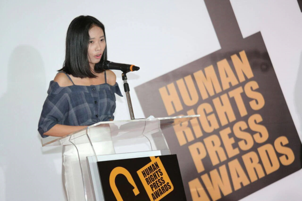 Eine Frau in einem schwarzen Kleid steht an einem Rednerpult mit einem Mikrofon und spricht ernst vor einem "Human Rights Press Awards"-Banner.