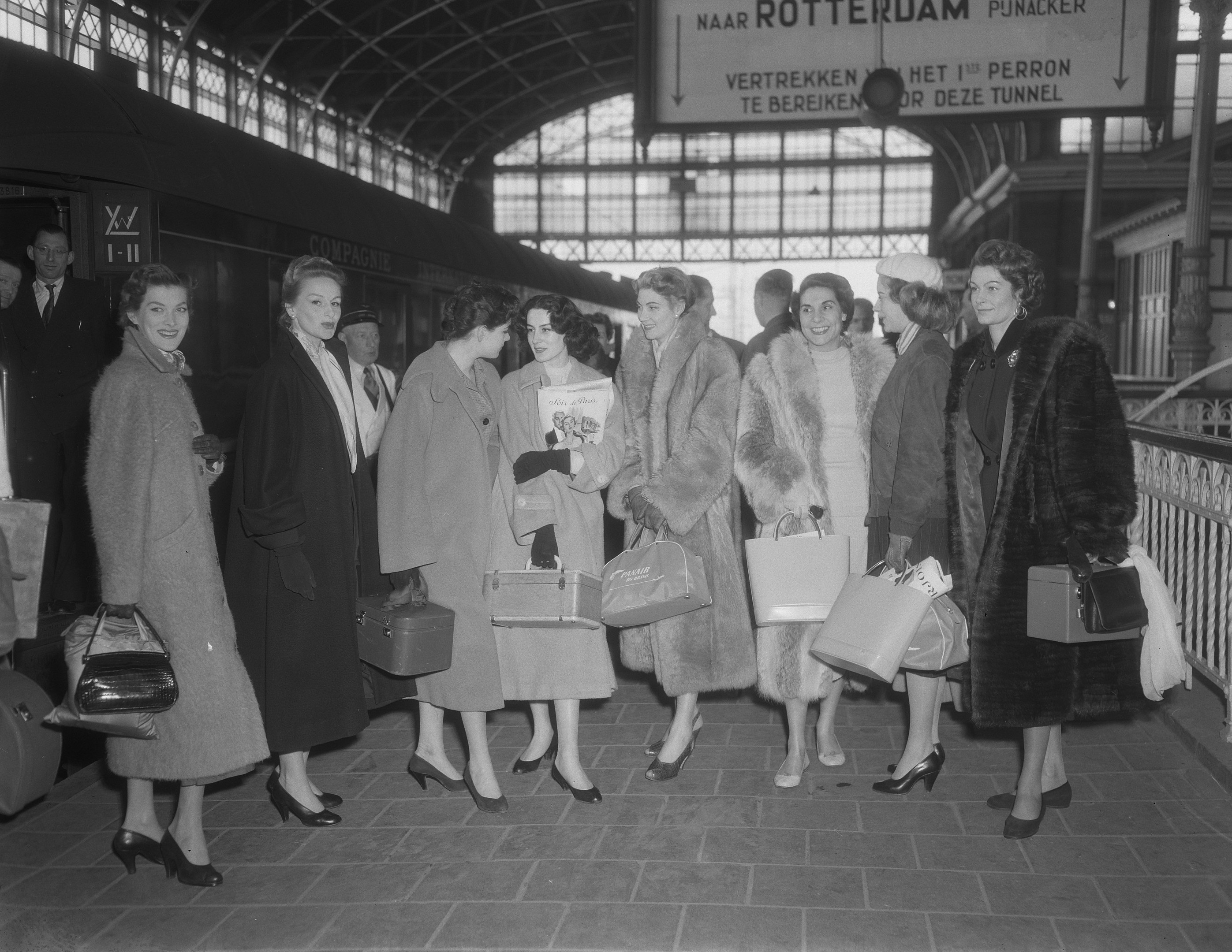 Schwarz-weißes Bild einer Gruppe von Frauen, die an einem Bahnhof stehen und jeweils eine Tasche halten, mit einem Zug auf der linken Seite und Geländern und einer Tafel auf der rechten Seite.