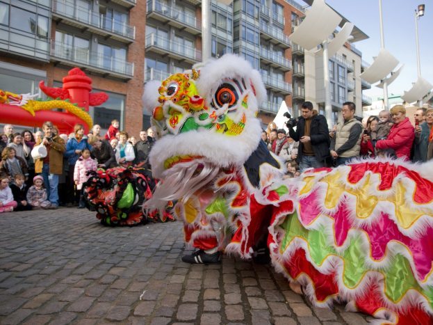 Ein lebendiges chinesisches Neujahrsfest in Amsterdam mit einer Löwendance-Vorführung vor einer Zuschauermenge, einige halten Kameras, vor dem Hintergrund von Gebäuden und einem klaren blauen Himmel.