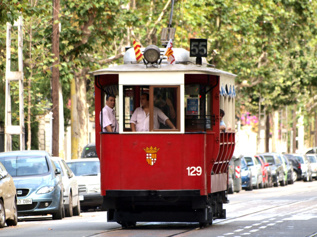 Rote Straßenbahn auf einer Stadtstraße mit Autos, Passagieren im Inneren und Bäumen und Strommasten im Hintergrund.
