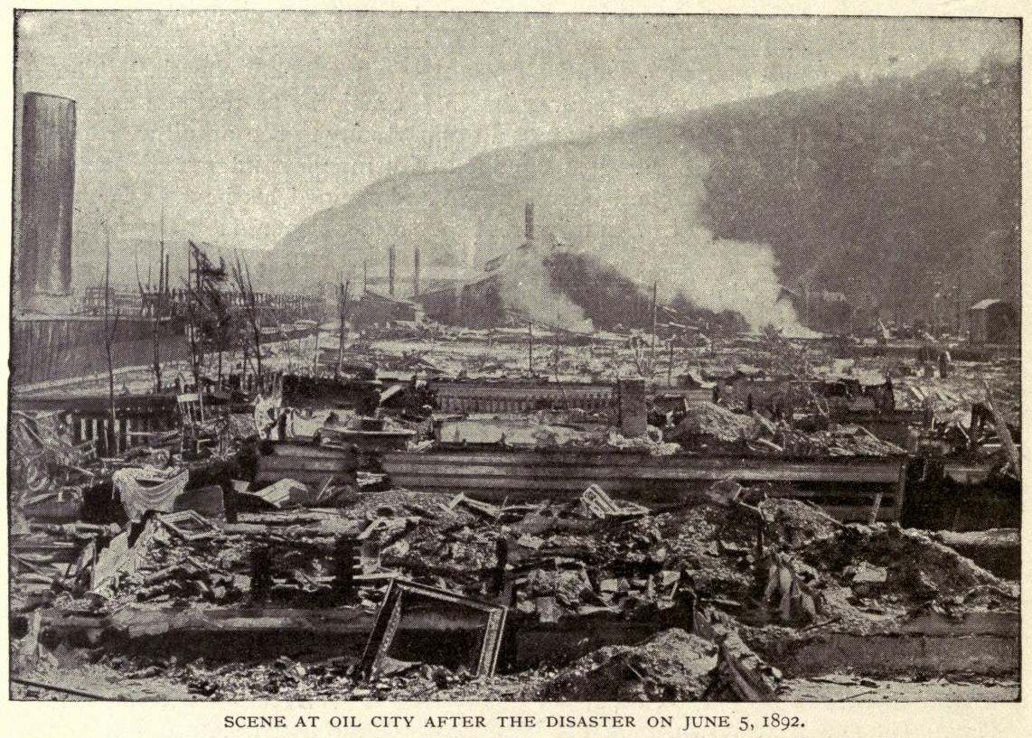 Schwarzes und weißes Foto einer Stadt nach einer Katastrophe am 5. Juni 1892, das zerstörte Gebäude, Strommasten, Rauch und einen Hügel im Hintergrund zeigt, mit Text unten.