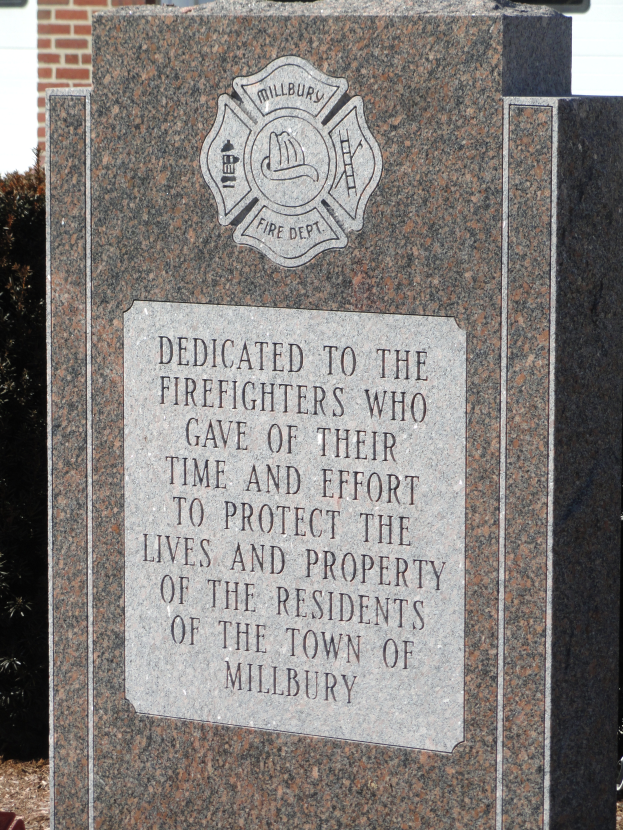 Ein Gedenkstein mit einer Plakette vor einem Backsteingebäude, umgeben von Pflanzen und Bäumen, gewidmet den Feuerwehrleuten von Millbury.