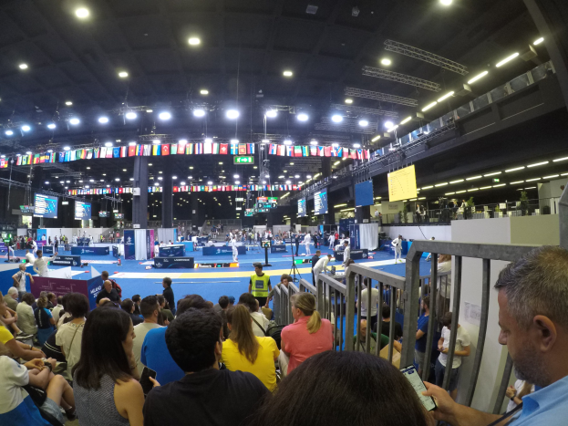 Große Menschenmenge sitzend und stehend in einer Indoor-Sportarena mit Bildschirmen, Fahnen und Deckenbeleuchtung im Hintergrund.