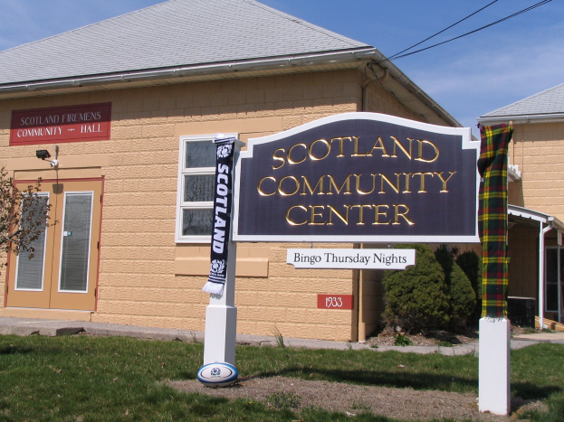Außenansicht des Scotland Community Centers mit einem Namensschild, Gras, einem Ball, Pflanzen, einem Baum, Stromleitungen und einem bewölkten Himmel.