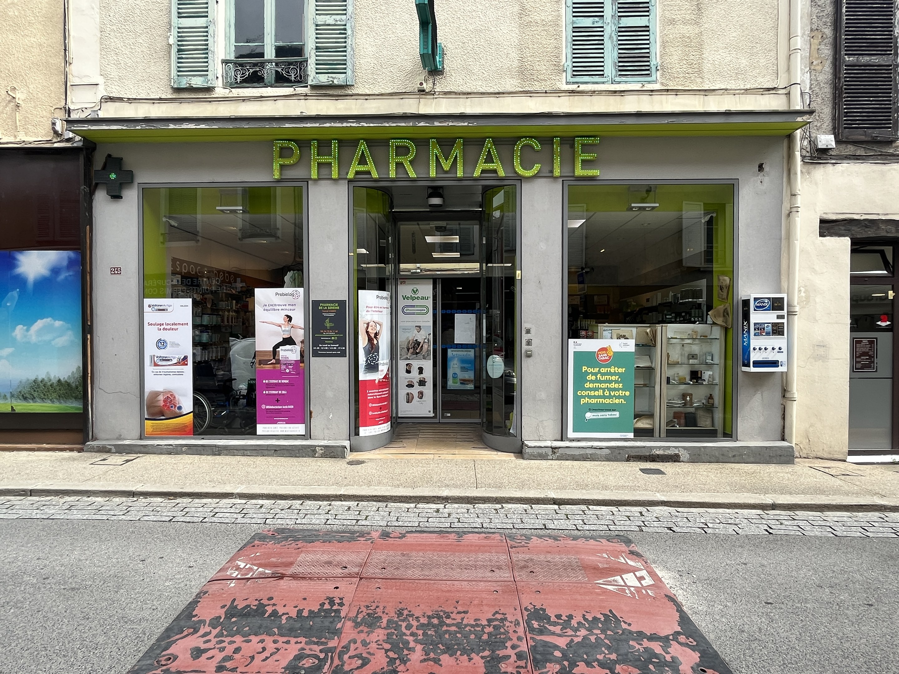 Apotheke mit Glaswänden und -türen, Plakate an den Wänden, an einer Straßenecke gelegen, mit einer Straße unten.