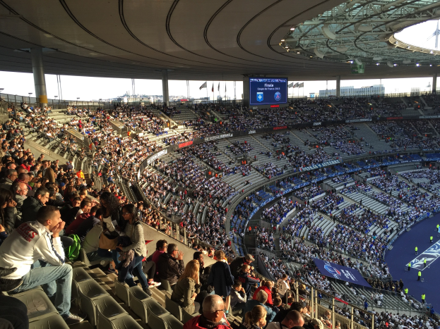 Eine große Menschenmenge in einem Stadion schaut ein Fußballspiel zu, mit einer Bühne, Fahnen und einem Bildschirm im Hintergrund, wahrscheinlich im Allianz Arena in München, Deutschland.