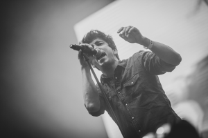 Ein Mann singt in ein Mikrofon auf einem Konzert, mit einem menschlichen Gesicht im Hintergrund, dargestellt in Schwarz-Weiß.