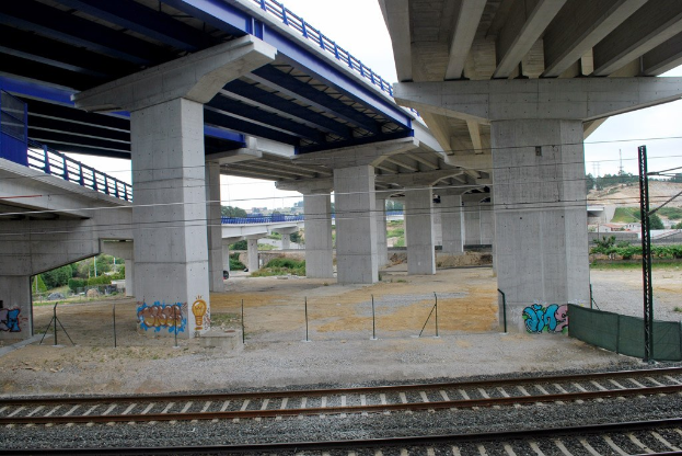 Eisenbahnschiene mit Graffiti unter einer Brücke, umgeben von Pfählen mit Drähten, Bäumen, Gebäuden und einem klaren blauen Himmel.