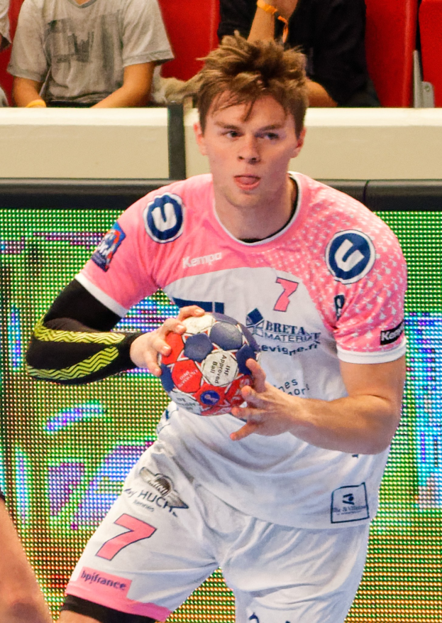 Ein Mann in einem rosa Hemd hält einen Fußball, mit der Hand eines anderen Menschen auf der linken Seite; im Hintergrund sitzen ein paar Menschen auf Stühlen, was auf ein Handballspiel hindeutet.