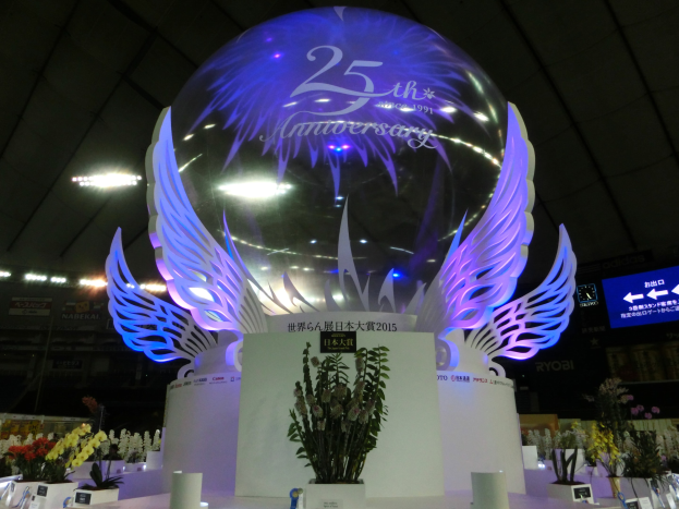 Ein großer Kristallball mit Flügeln oben drauf, umgeben von Blumenbouquets, Namensschildern und anderen Gegenständen auf dem Boden, mit Tafeln, Lichtern und einem Bildschirm im Hintergrund bei einer festlichen 25-jährigen Jubiläumsfeier.