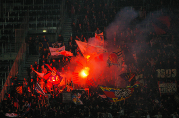 Eine große Menschenmenge in einem Stadion mit Fahnen, Bannern und einem zentralen Feuer, beleuchtet von Lichtern und umgeben von Stufen und Geländern.