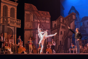 Ein Mann und eine Frau tanzen auf einer Bühne im Royal Opera House in London, mit einer Menschenmenge im Vordergrund und einem Bildschirm mit Gebäuden im Hintergrund.