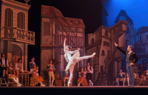 Ein Mann und eine Frau tanzen auf einer Bühne im Royal Opera House in London, mit einer Menschenmenge im Vordergrund und einem Bildschirm mit Gebäuden im Hintergrund.