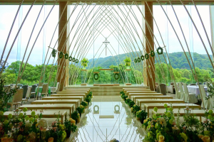 Eine Hochzeitszeremonie in einem großen Gebäude mit Tischen und Stühlen in einem Halbkreis angeordnet, dekoriert mit Blumenbouquets, mit Bäumen, Hügeln und einem klaren blauen Himmel im Hintergrund.
