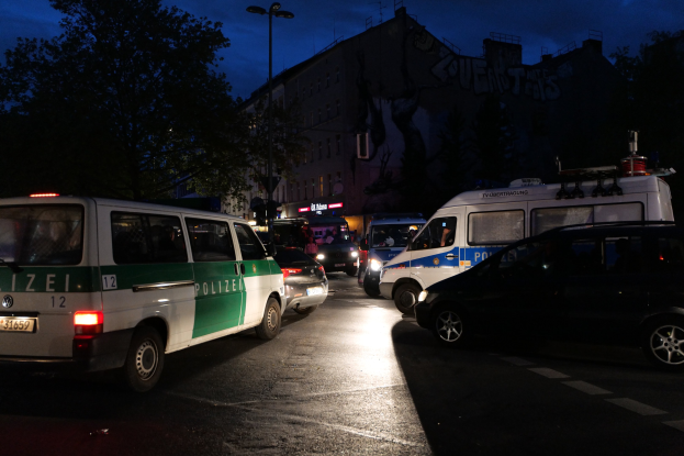 Eine Gruppe von Polizeiwagen, darunter ein Van, ein Auto und ein Rad, parkt nachts am Straßenrand mit Gebäuden, Bäumen, Laternenmasten und einem Sternenhimmel im Hintergrund.