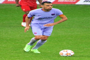 Ein Fußballspieler in einem Sportoutfit und -schuhen ist im Einsatz auf dem Feld, wobei ein Schild mit Text im Hintergrund sichtbar ist.