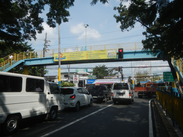 Verkehrsreiche Straße mit Fahrzeugen, eine Brücke mit Geländern und Treppen, Laternen, Verkehrsampeln, Schilder, Bäume, Gebäude und ein bewölkter Himmel.