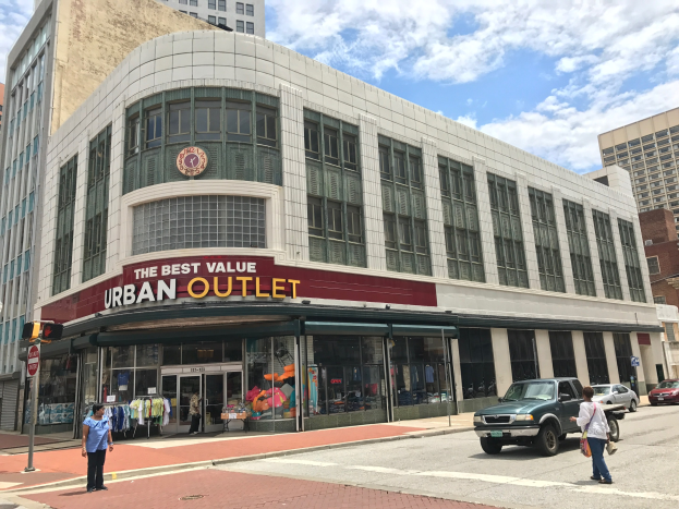 Eine belebte Stadtstraße mit fahrenden Fahrzeugen und Fußgängern auf dem Gehweg, hohe Glasgebäude im Hintergrund und ein sichtbares "The Best Value Urban Outlet"-Schild unter einem bewölkten Himmel.