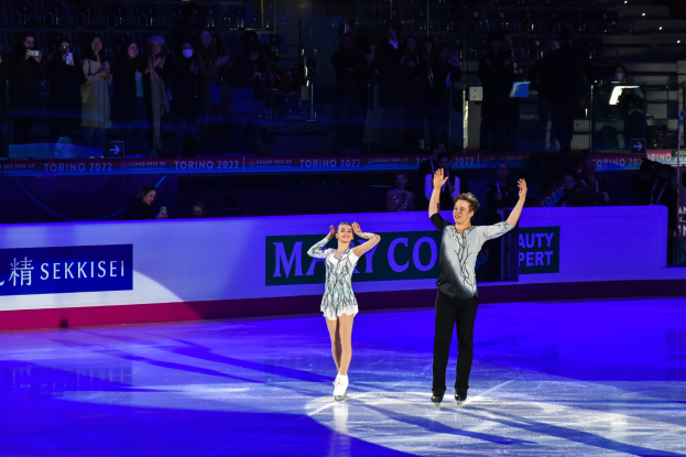 Zwei Personen beim Eislaufen auf einer Eisbahn vor Publikum mit einigen Fotografen, unter heller Beleuchtung mit einem Textschild im Hintergrund.