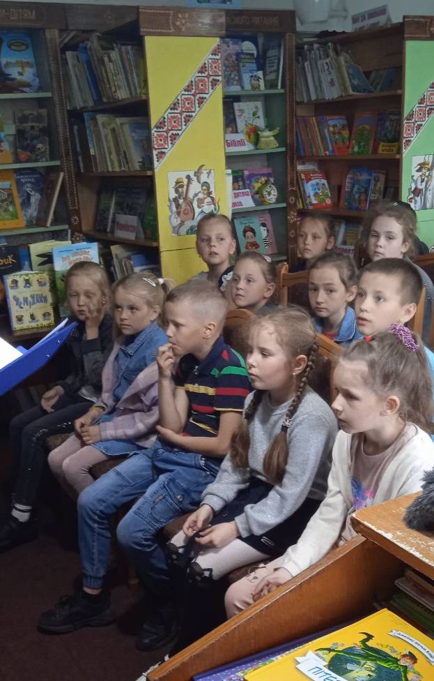 Eine Gruppe von Kindern sitzt in einer Bibliothek umgeben von Bücherregalen, wobei einige Bücher an Tischen auf beiden Seiten des Bildes lesen.