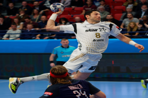Zwei Männer beim Handballspielen auf einem Platz, einer springt mit einem Ball, der andere steht in der Mitte, mit einer Zuschauermenge auf Stühlen und einem blauen Schild im Hintergrund mit der Aufschrift "Futsal-Weltmeisterschaft 2015".