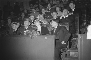 Ein Schwarz-Weiß-Foto einer Menschenmenge in einem Stadion, mit einigen auf Bänken sitzend und anderen stehend, die einer Tribüne mit einem Blumenstrauß und einer Wand im Hintergrund zugewandt sind.