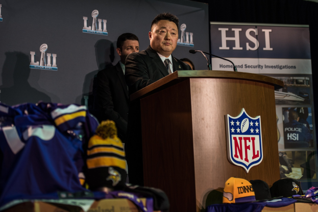 Ein Mann in einem schwarzen Blazer, einem weißen Hemd und einer schwarzen Krawatte spricht an einem Podium mit einem Mikrofon und Logo vor einer Menge, mit blauer und gelber Merchandise zu seiner Linken und einem "Super Bowl LII"-Banner im Hintergrund.