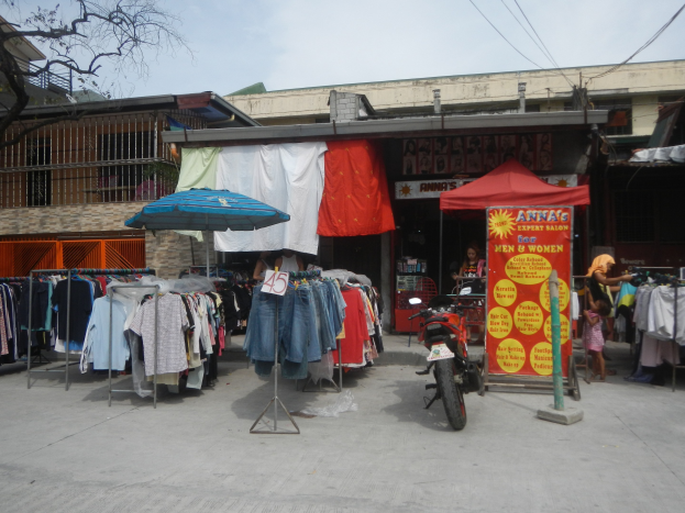 Eine Straßenszene mit einem vor einem Bekleidungsgeschäft geparkten Motorrad, das Kleidungsstücke auf Bügeln, einen Schirm und eine nahestehende Person zeigt, mit Gebäuden, einem Baum und dem Himmel im Hintergrund.