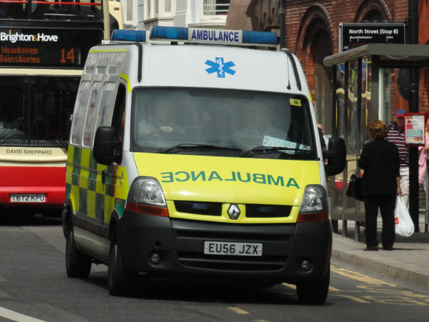 Rettungswagen fährt auf einer Straße neben einem roten Doppeldeckerbus mit Fußgängern auf dem Gehweg und Gebäuden im Hintergrund.