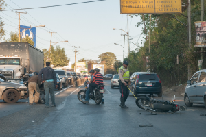 Eine Gruppe von Menschen steht um ein verunglücktes Motorrad am Straßenrand mit mehreren Fahrzeugen, darunter ein Lastwagen, und einem Hintergrund aus Bäumen, Pfählen, Lampen, Schildern und Himmel.