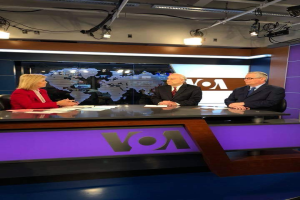 Drei Personen, zwei Männer in Blazern und Krawatten und eine Frau in einem roten Kleid sitzen an einem Schreibtisch mit Papieren und anderen Gegenständen und führen eine Diskussion vor einem Fernseher mit einem Bildschirm und Lichtern im Hintergrund.